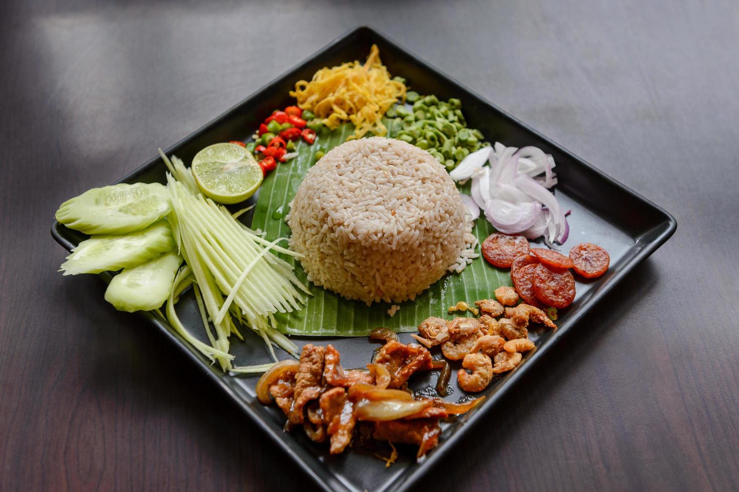 thailändisches Essen - Reis gemischt mit Garnelenpaste, Kao cluk ka pi mit Beilage als Mango, Zitrone, Chili, Gurke, Rührei, Kuherbse, Schalotten, chinesischer Wurst, getrockneten Garnelen und Schweinefleisch foto