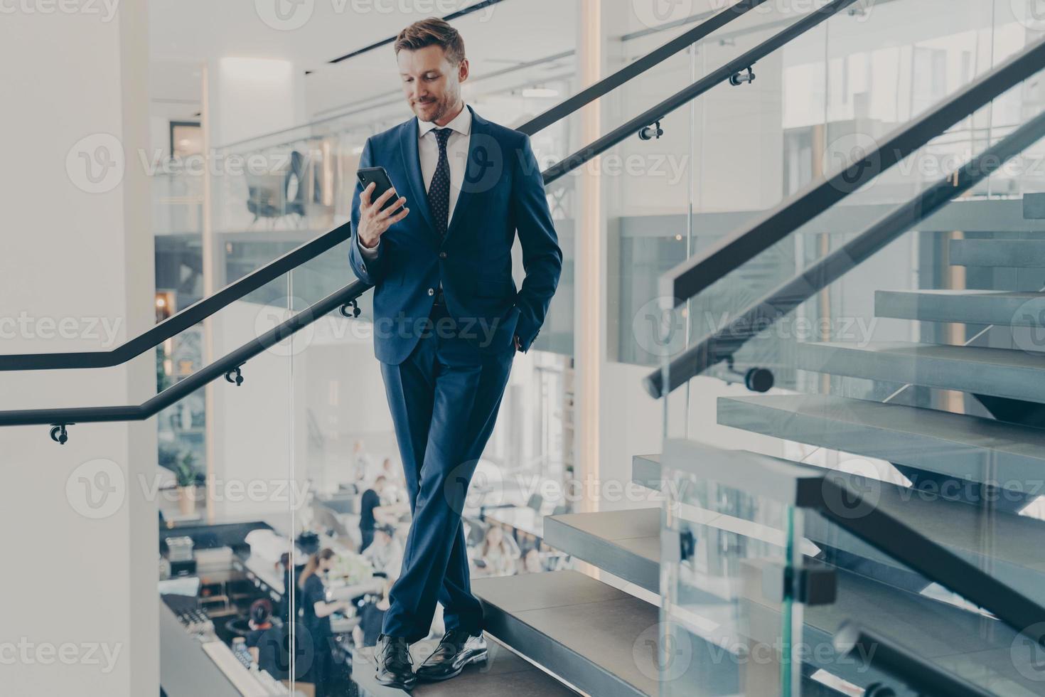 Atemberaubender Unternehmer in formellem, stilvollem Anzug, der mit Smartphone auf der Treppe in der Bürolobby steht foto