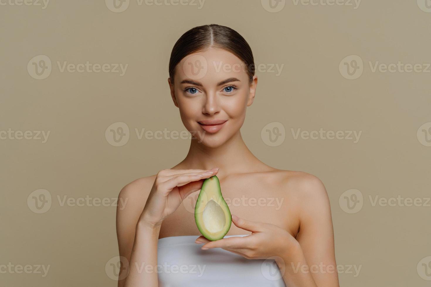 Hygienekonzept für Hautpflege und Gesichtsbehandlungen. innenaufnahme eines erfreuten weiblichen models hält die hälfte der avocado, die in ein badetuch gewickelt wird, das ein schönheitsprodukt verwenden wird, nährt die haut, die über einer beigen wand isoliert ist foto