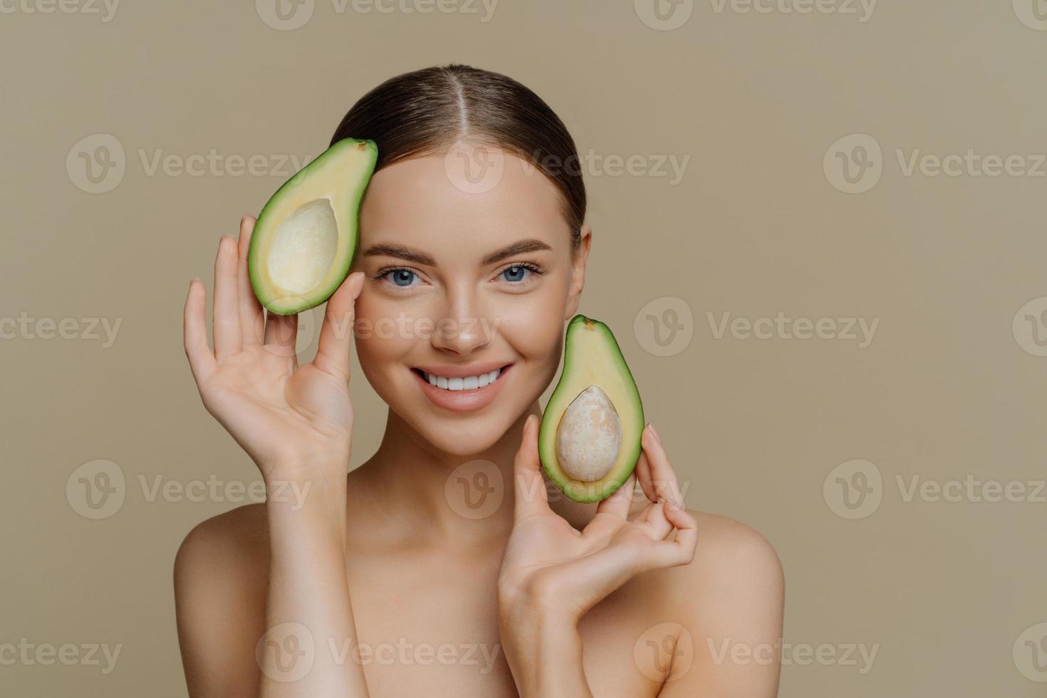 Gesichtsbehandlung mit natürlichen Vitaminen. zufriedene brünette frau lächelt sanft hält hälften von avocado schaut direkt auf die kamera steht oben ohne drinnen hat gesunde haut isoliert über beiger wand. foto