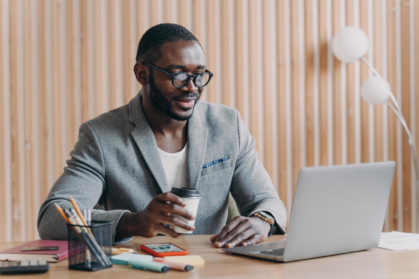 junges lächelndes afroamerikanisches geschäft in brille, die mit kaffeetasse am arbeitsplatz im büro sitzt foto