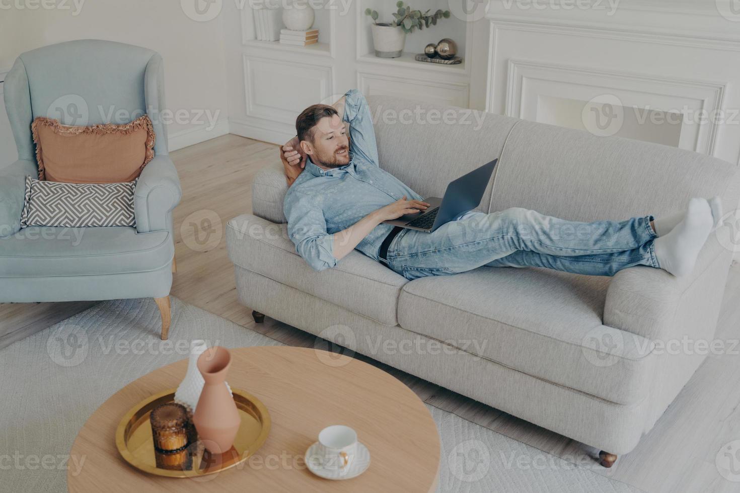 Junger Geschäftsmann, der zu Hause aus der Ferne arbeitet, während er auf der Couch liegt foto