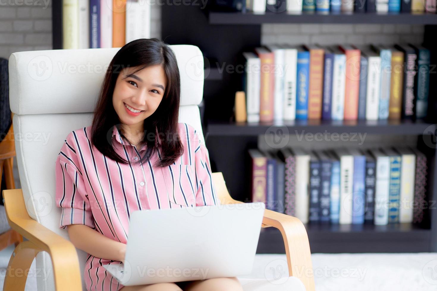 schöne asiatische junge frau des porträts, die online-laptop mit lächeln und glücklichem sitzen auf stuhl im wohnzimmer, mädchen mit notebook-computer mit verbindung zum internet, geschäfts- und erfolgskonzept arbeitet. foto