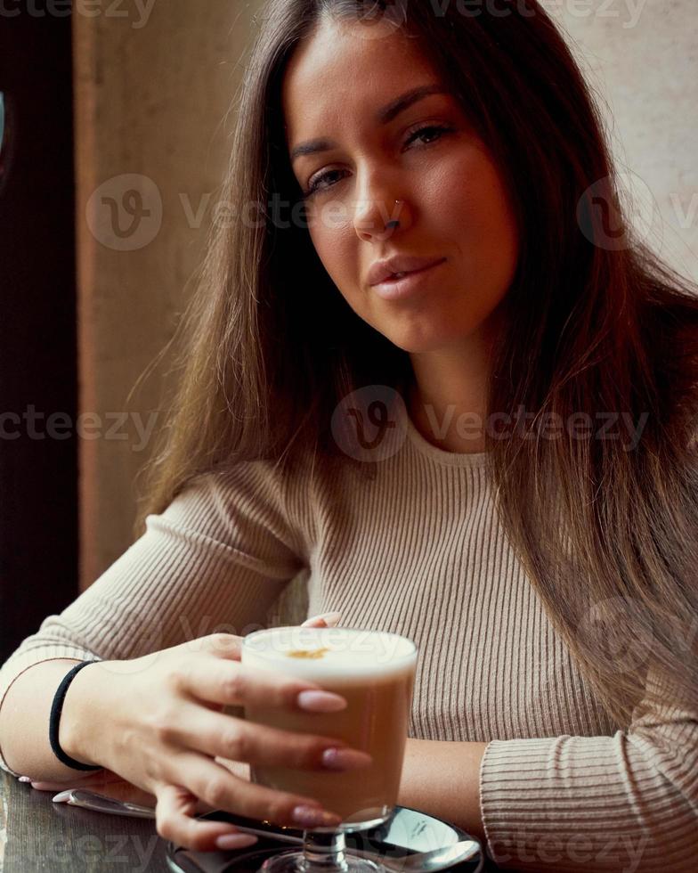 schönes nachdenkliches glückliches mädchen, das in den weihnachtsferien im café sitzt, lächelt und träumt. Brünette Frau mit langen Haaren trinkt Cappuccino, Latte und schaut in die Kamera foto