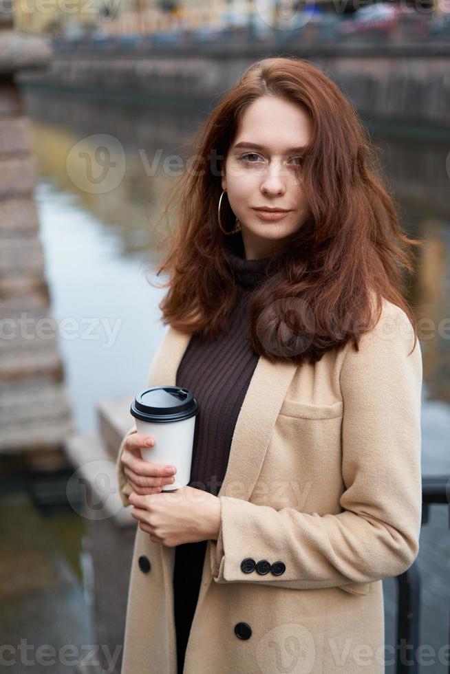 schönes ernstes stilvolles modisches mädchen, das tasse kaffee in den händen hält gehende straße von st. Petersburg im Stadtzentrum. charmante nachdenkliche frau mit langen dunklen haaren schaut in die kamera foto