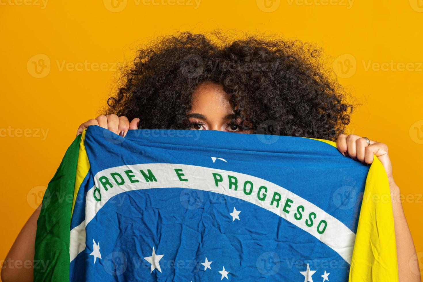 geheimnisvoller schwarzer frauenfan, der eine brasilianische flagge in deinem gesicht hält. Brasilien-Farben im Hintergrund, grün, blau und gelb. Wahlen, Fußball oder Politik. foto