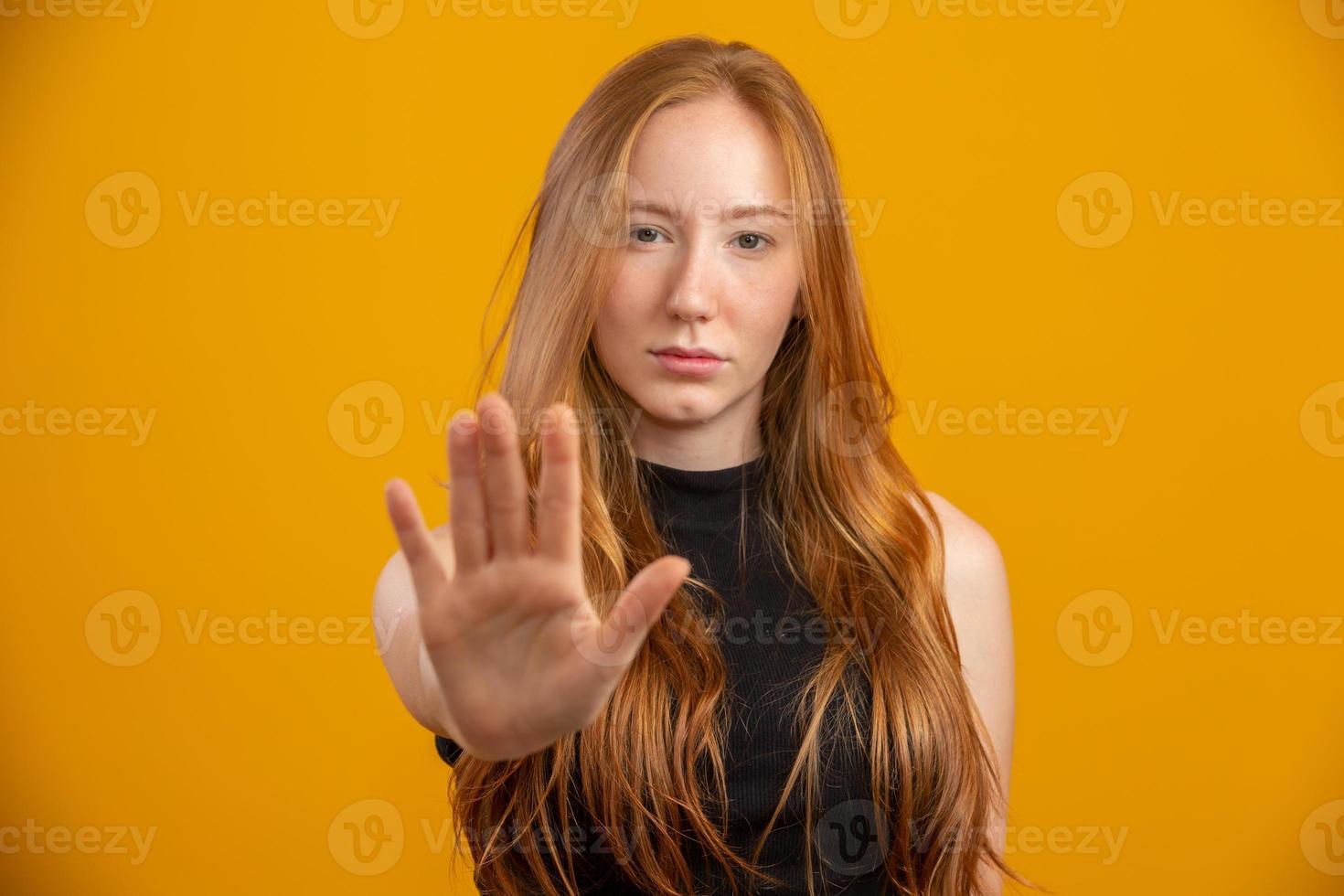 Schöne rothaarige Frau, die über isoliertem gelbem Hintergrund steht und mit offener Hand ein Stoppschild mit ernstem und selbstbewusstem Ausdruck macht, Verteidigungsgeste. Keine Gewalt mehr gegen Frauen. Missbrauch. foto