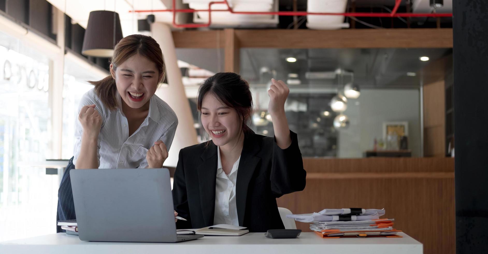zwei junge asiatische geschäftsfrauen zeigen einen freudigen ausdruck des erfolgs bei der arbeit und lächeln glücklich mit einem laptop in einem modernen büro. foto