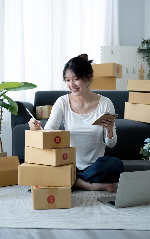 lächelnde junge asiatische geschäftsinhaberin bereiten paketkasten vor und prüfen online-bestellungen von produkten für die lieferung an den kunden auf einem laptop-computer. Online-Shopping-Konzept. foto