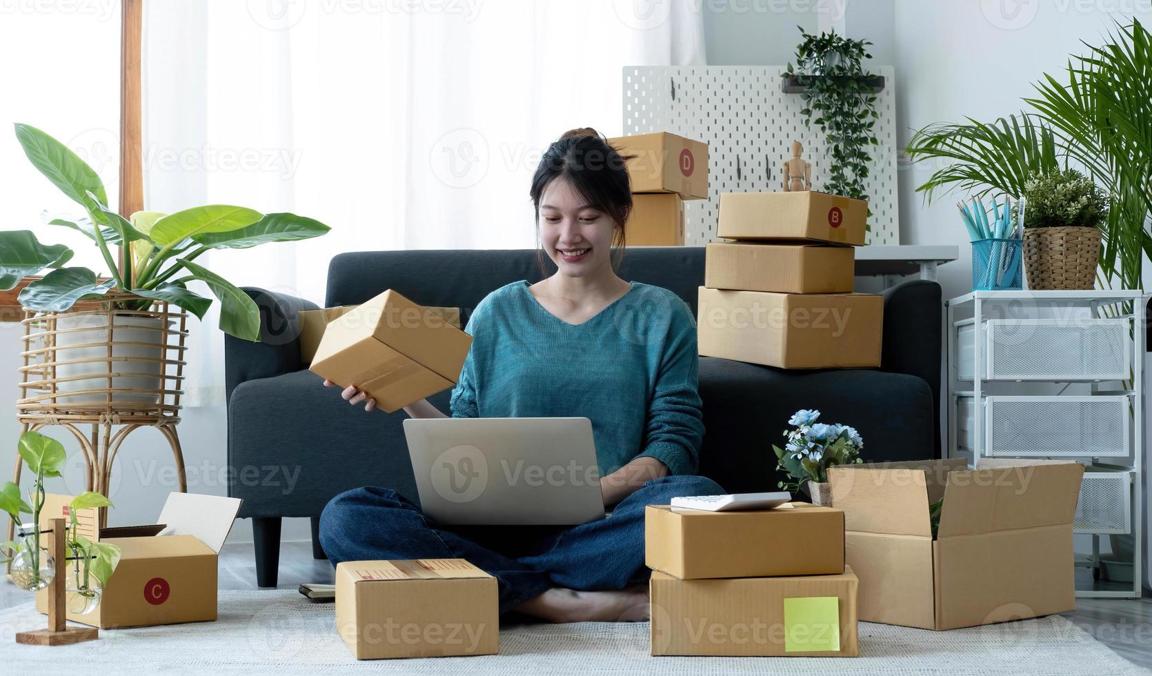 Fröhliche junge Asiatin Startup Kleinunternehmen freiberuflich tätig, die Paketbox und Computer-Laptop hält und auf dem Boden sitzt, Online-Marketing-Verpackungsbox-Lieferkonzept foto