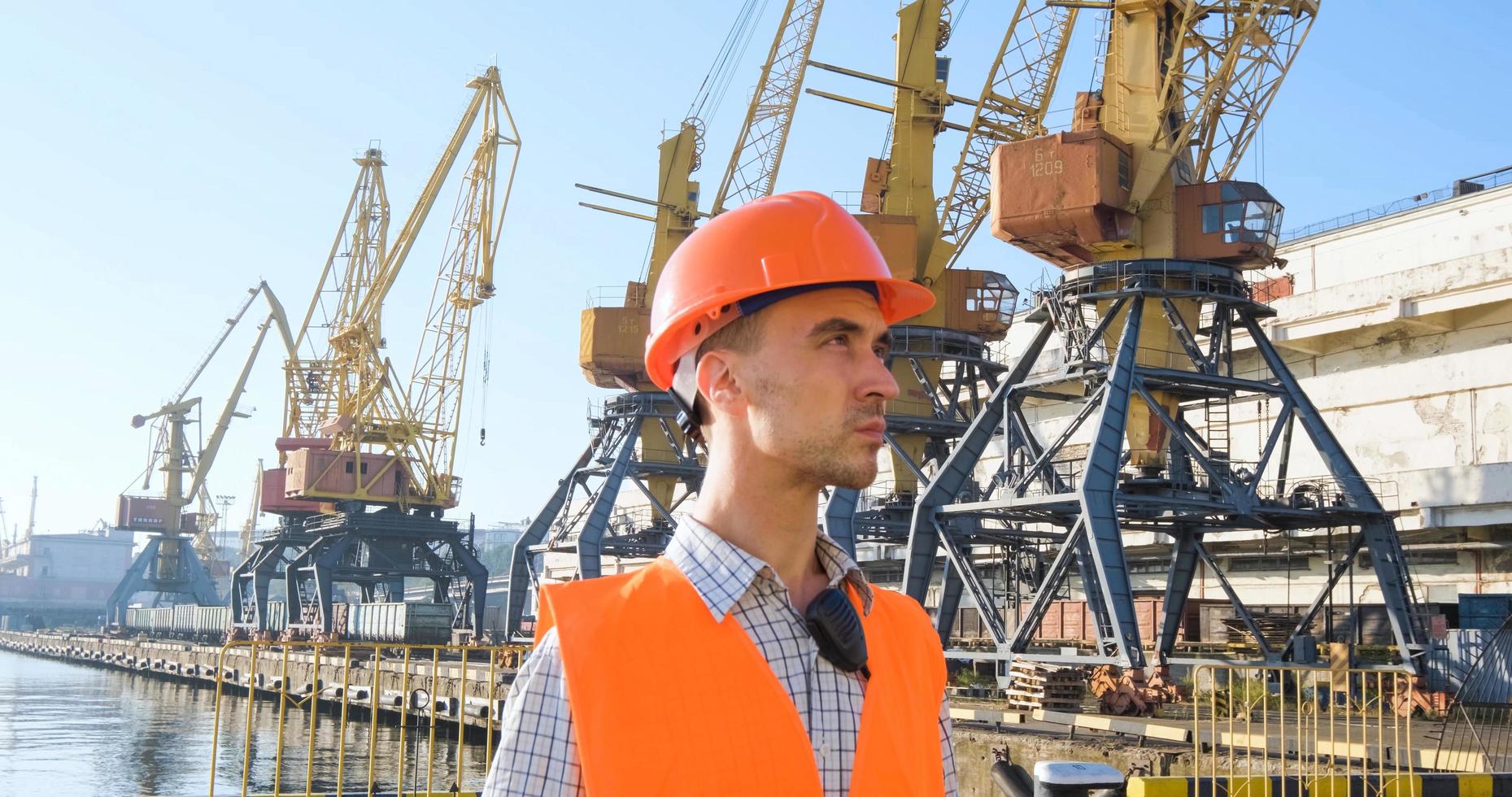männlicher arbeiter des seehafens in orangefarbenem helm und sicherheitswesten, kranen und meereshintergrund foto