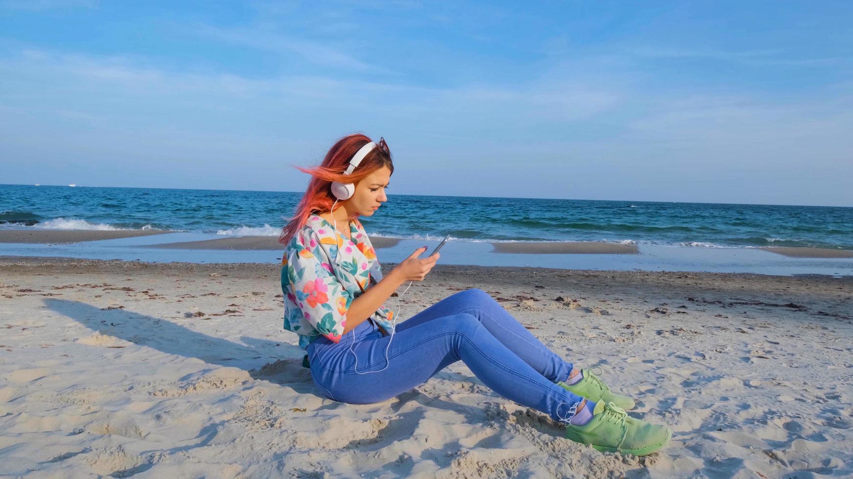 junge frau mit bunten haaren geht am sommerstrand spazieren und hört musik mit kopfhörern foto