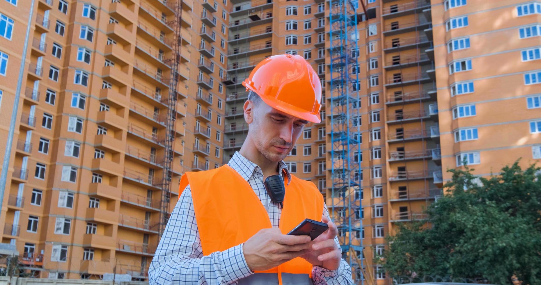 Porträt des Baufachmanns in orangefarbenem Helm und Sicherheitsweste gegen großes Gebäude foto