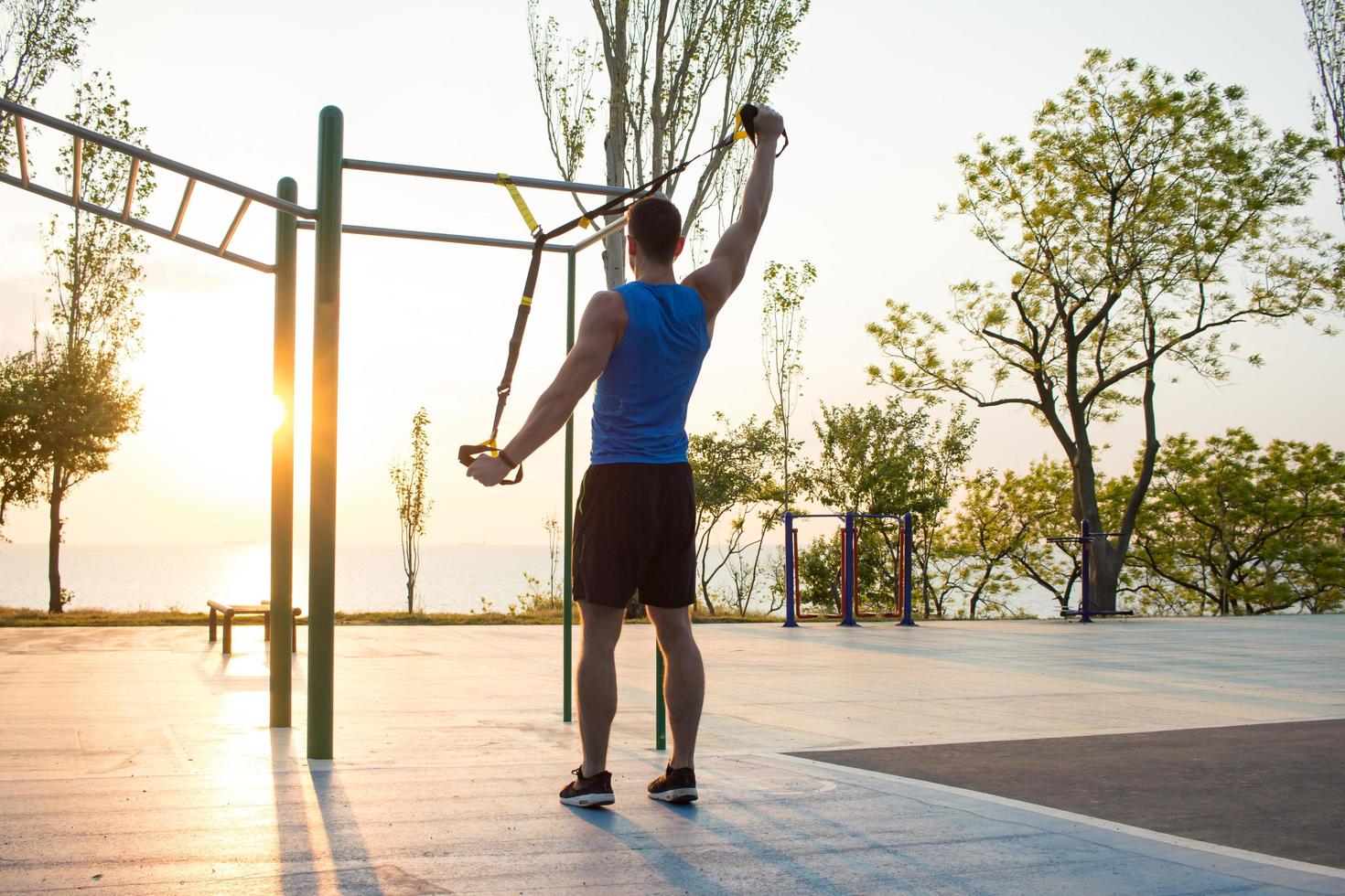 Training mit Aufhängegurten im Fitnessstudio im Freien, starkes Manntraining am frühen Morgen im Park, Sonnenaufgang oder Sonnenuntergang im Meereshintergrund foto