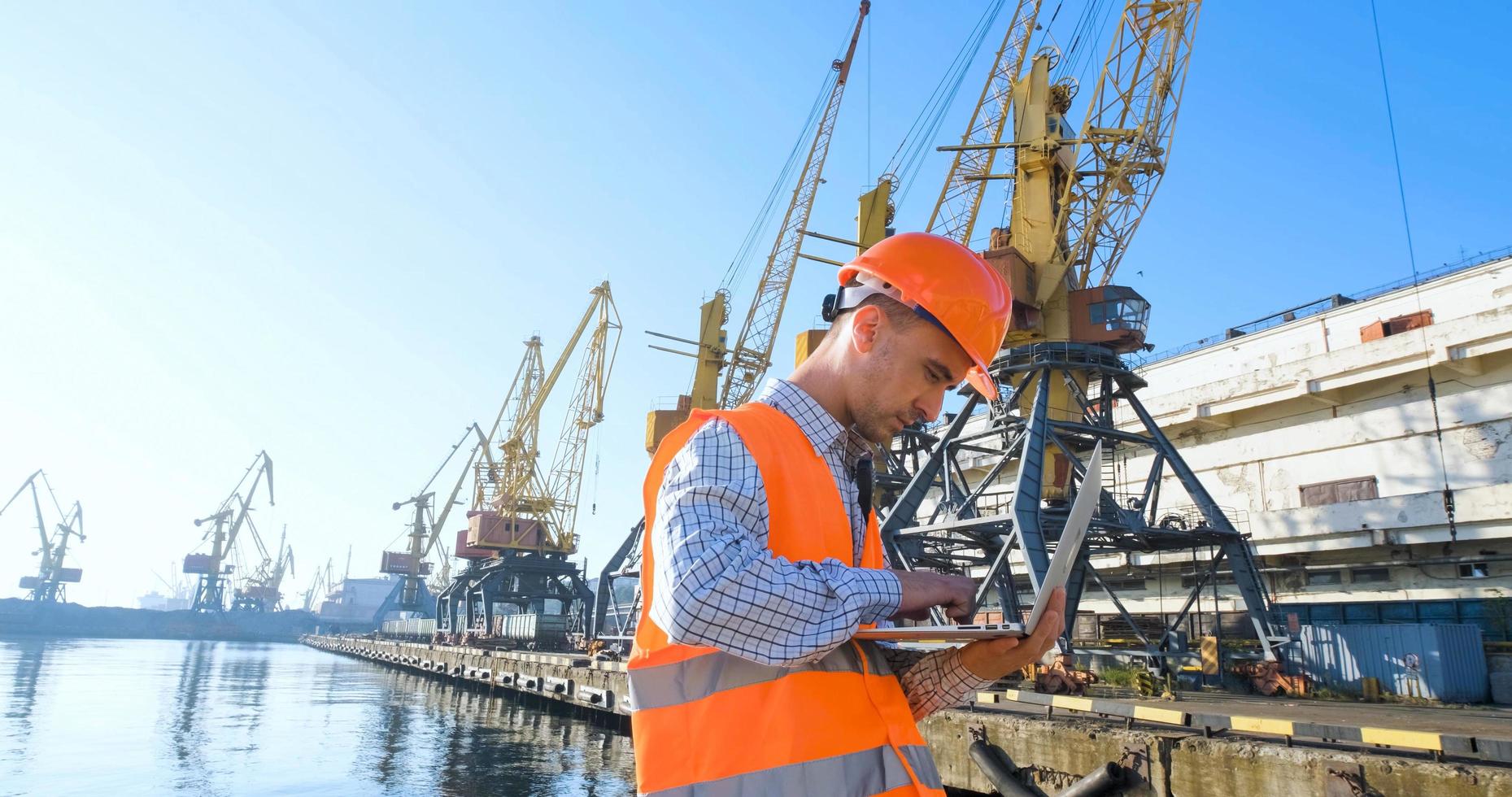 männlicher arbeiter des seehafens in orangefarbenem helm und sicherheitswesten, kranen und meereshintergrund foto