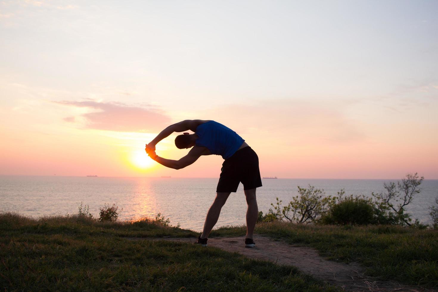 Fröhlicher fitter Läufer, der Sonnenaufgang oder Sonnenuntergang mit erhobenen Fäusten beobachtet, junger Athlet auf Gras während des Sonnenaufgangs im Meer foto