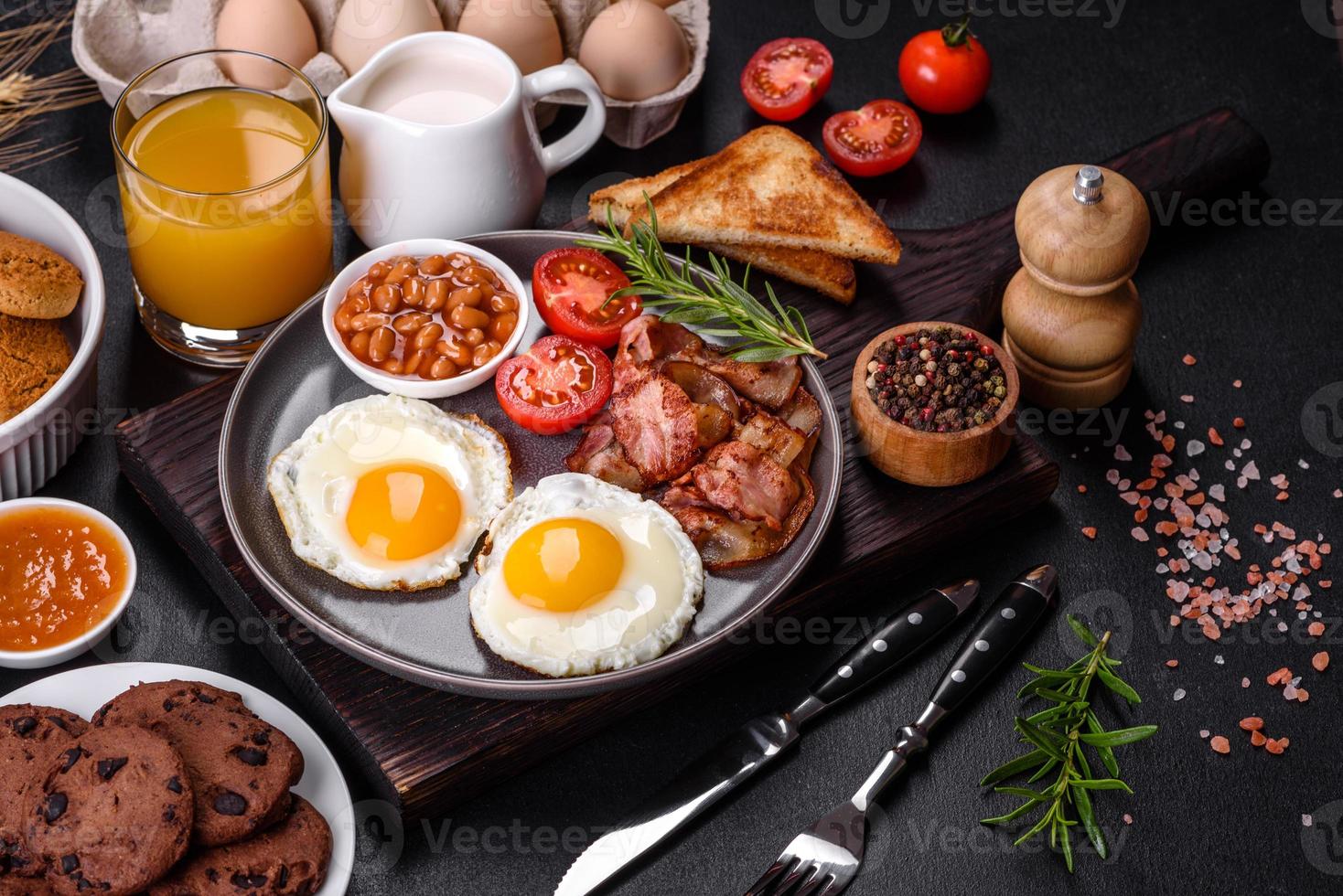 Ein köstliches, nahrhaftes, gesundes Frühstück mit Spiegeleiern, Speck, Bohnen, einem Glas Saft, Haferkeksen, Milch und Marmelade foto