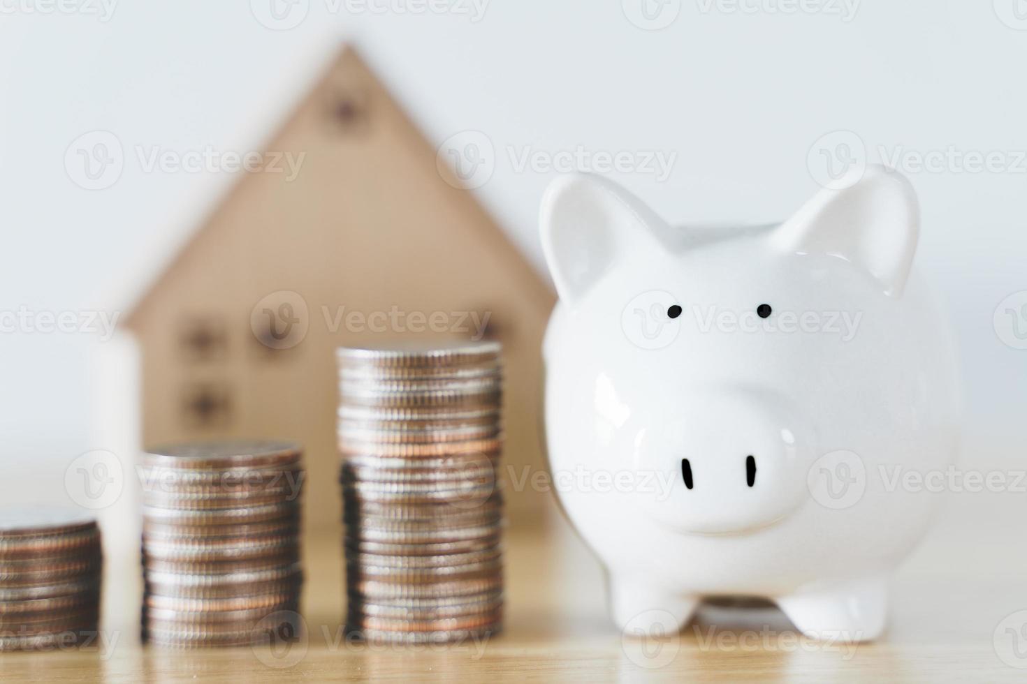 Holzhaus mit weißem Sparschwein und Stapeln von Münzen auf Holztisch. Geld sparen für den Kauf eines Hauses, Finanzierungskonzept für Wohnungsbaudarlehen. foto