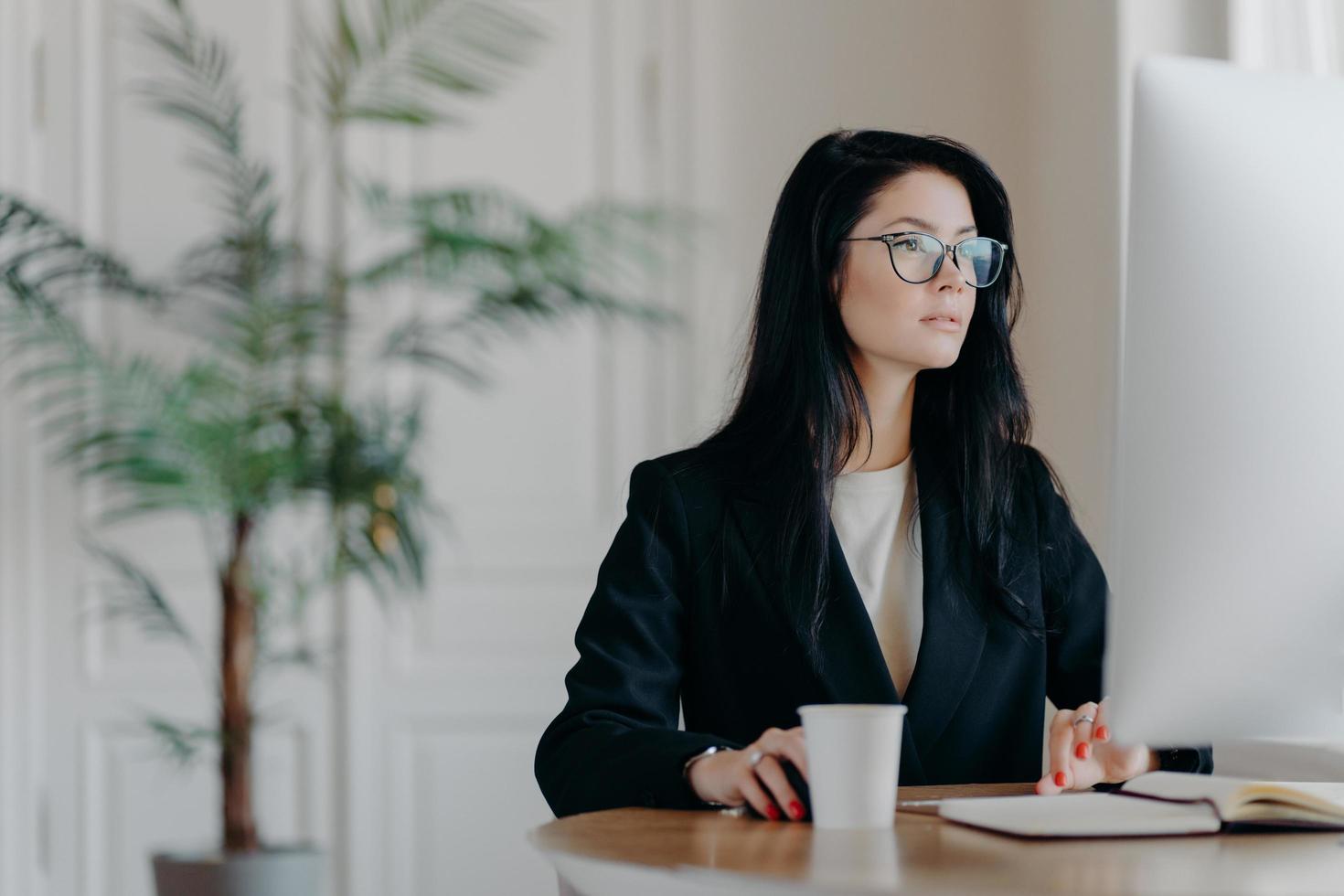 konzentrierte unternehmerin trägt ein elegantes schwarzes kostüm, arbeitet am computer, sitzt am tisch an einem gemütlichen arbeitsplatz trägt eine durchsichtige brille, surft im internet und lässt das geschäftsprojekt über die strategie nachdenken foto