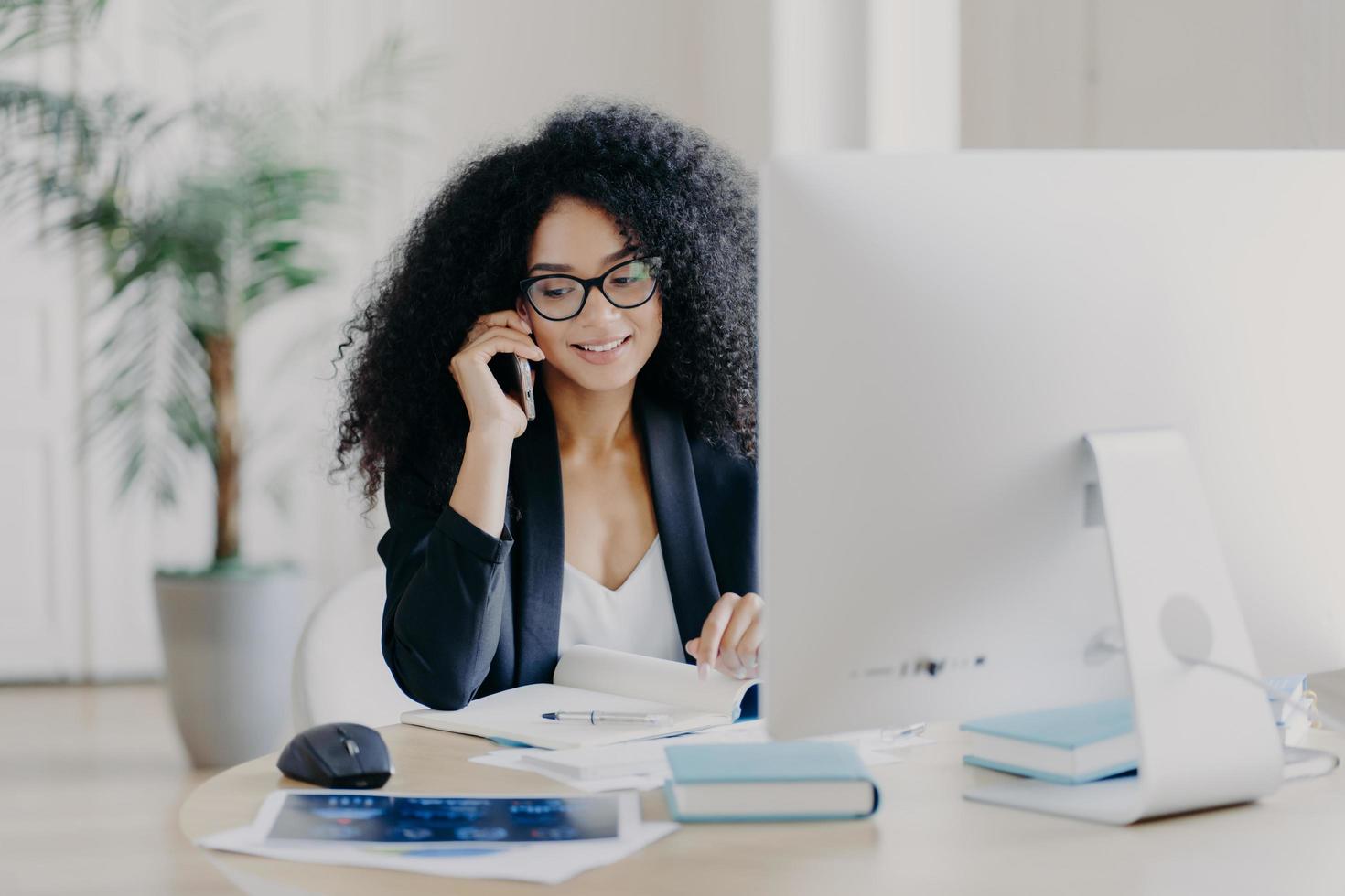 Foto einer zufriedenen Afroamerikanerin ruft per Smartphone an, macht sich Notizen im Notizblock, ist am Arbeitsprozess beteiligt, sitzt am Schreibtisch mit Computer, trägt eine Brille, formelle Kleidung, hat Telefonkommunikation