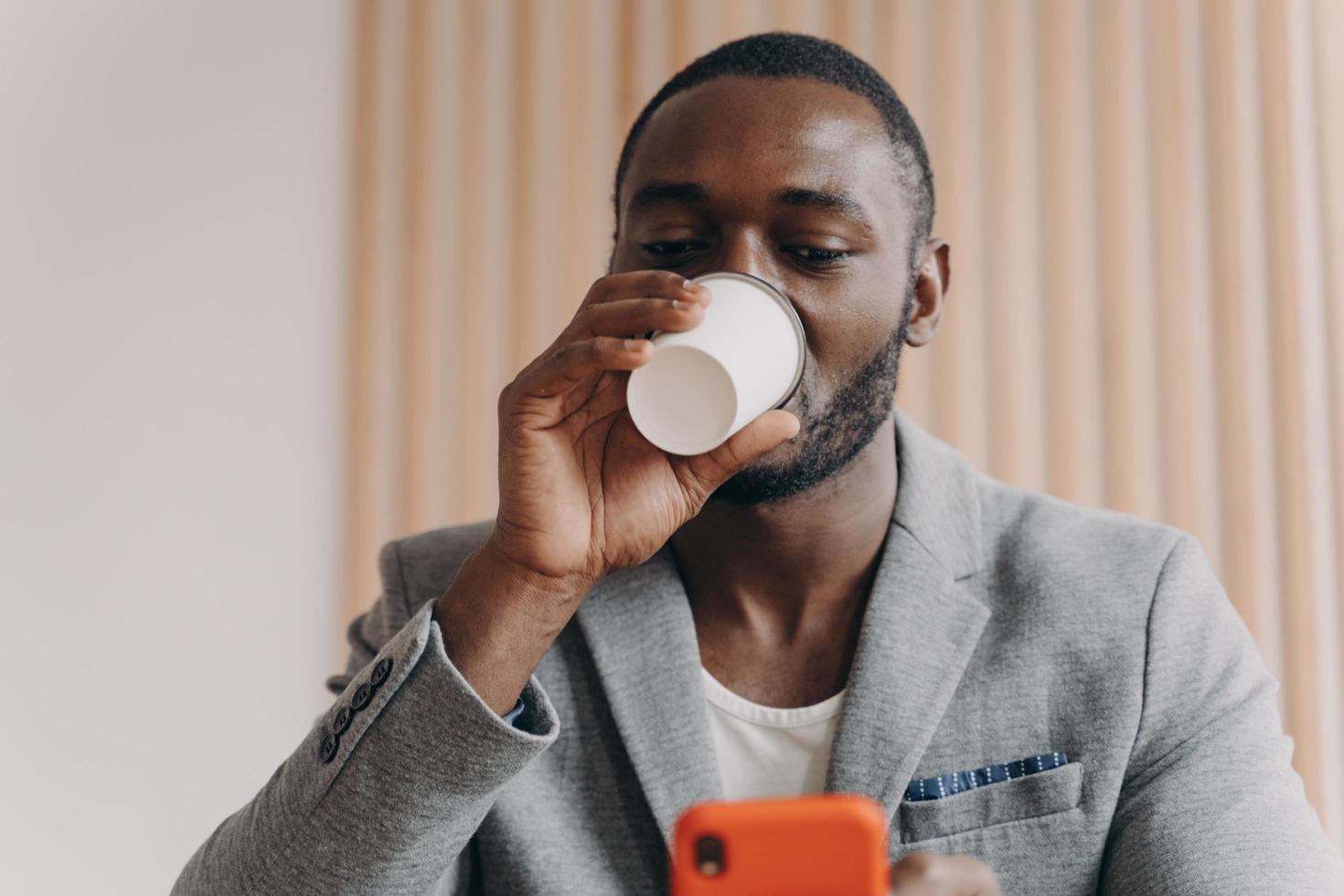 Eleganter afrikanischer Ethnizitätsgeschäftsmann, der Kaffee zum Mitnehmen trinkt, während er auf dem Handybildschirm schaut foto