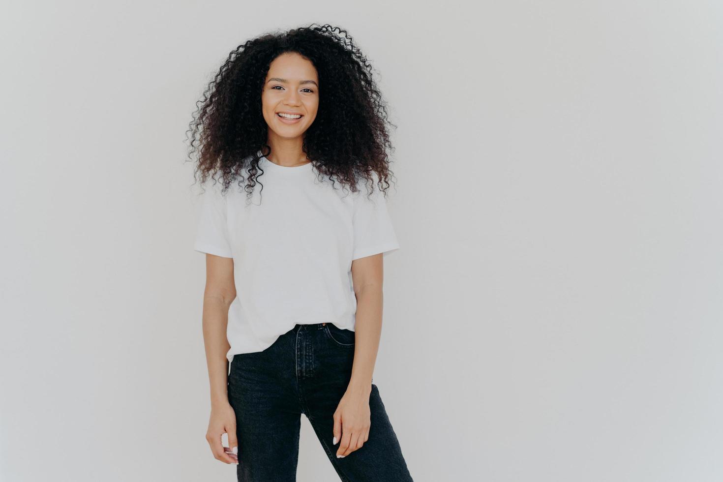 foto einer lächelnden frau mit lockigem dunklem haar, hat eine schlanke figur, trägt ein weißes t-shirt und schwarze jeans, hat einen fröhlichen gesichtsausdruck, modelle vor weißem hintergrund, kopierraum für ihre werbeinhalte