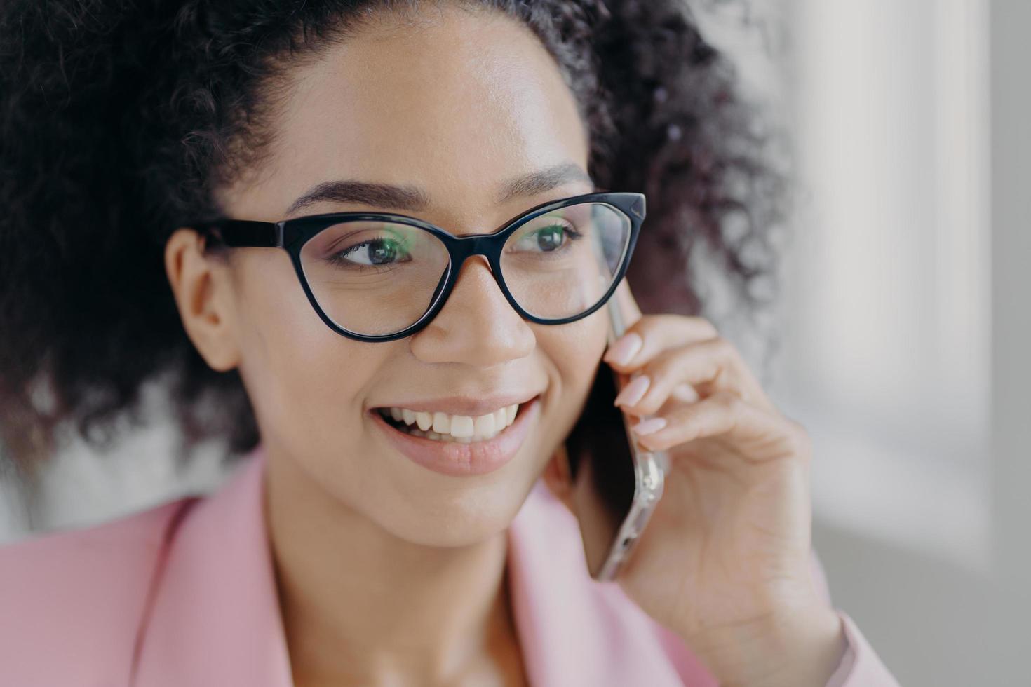 kopfschuss einer schönen lächelnden dunkelhäutigen frau trägt eine optische brille, hält ein modernes handy in der hand, hat einen gepflegten teint, spricht über zukünftige investitionen mit einem geschäftspartner, schaut zur seite. foto