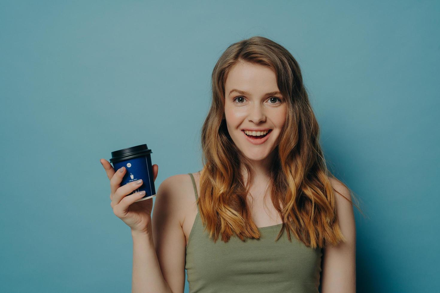 Fröhliche junge mädchenhafte Frau, die sich sorglos fühlt, eine Einweg-Kaffeetasse hält und in die Kamera lächelt foto