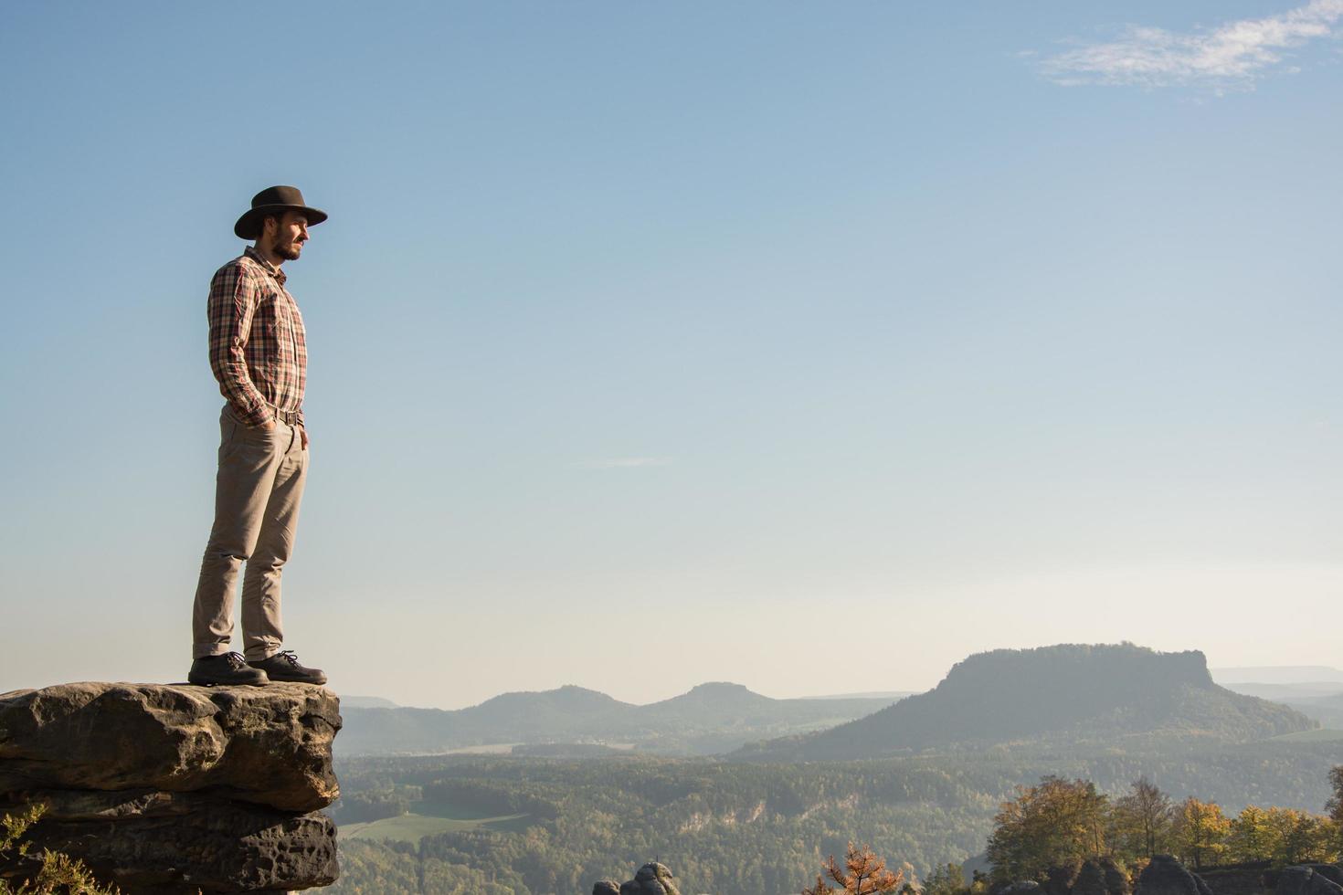 junger bärtiger männlicher Reisender mit Hut steht an sonnigen Tagen auf der Klippe foto