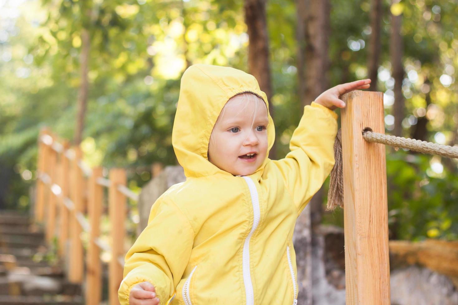 lustiges zweijähriges mädchen im gelben regenmantel spazieren im park am sonnigen tag foto