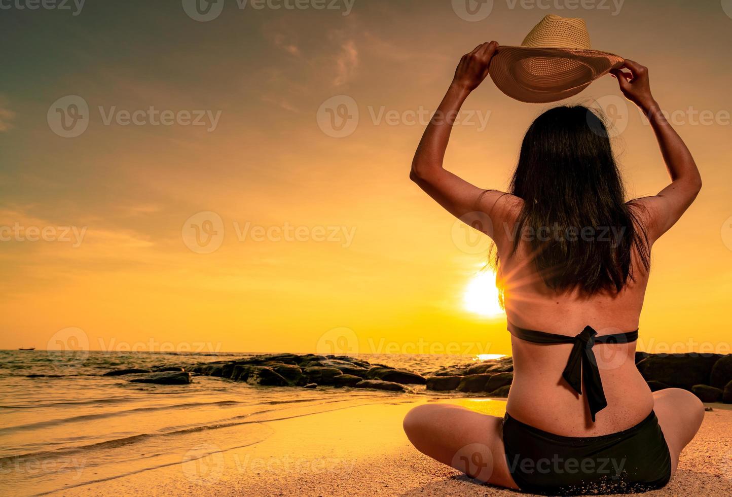 Rückansicht der dicken asiatischen Frau mit Bikini und Strohhut in der Hand sitzen am Sandstrand entspannen und Urlaub am tropischen Paradiesstrand genießen. Frau sieht schönen Sonnenuntergang. Sommerurlaub. sommergefühl. foto