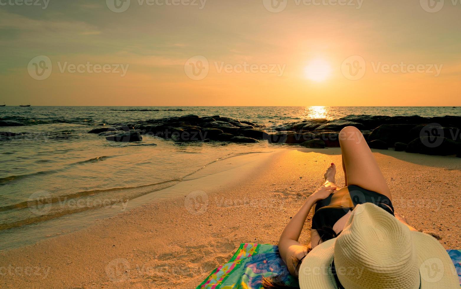 Frau, die sich bei Sonnenaufgang am Sandstrand hinlegt. Frau mit Strohhut beim Sonnenbaden am tropischen Paradiesstrand mit wunderschönem Sonnenaufgangshimmel. Sommerurlaub. Mädchen tragen schwarzen Badeanzug schlafen am Sandstrand. foto