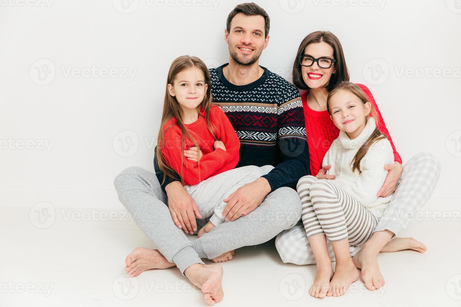 Foto der glücklichen einvernehmlichen Familie posieren alle zusammen vor weißem Hintergrund. freundlich aussehende mutter, vater und ihre beiden töchter sitzen auf dem boden, haben freudige gesichtsausdrücke. zusammengehörigkeitskonzept