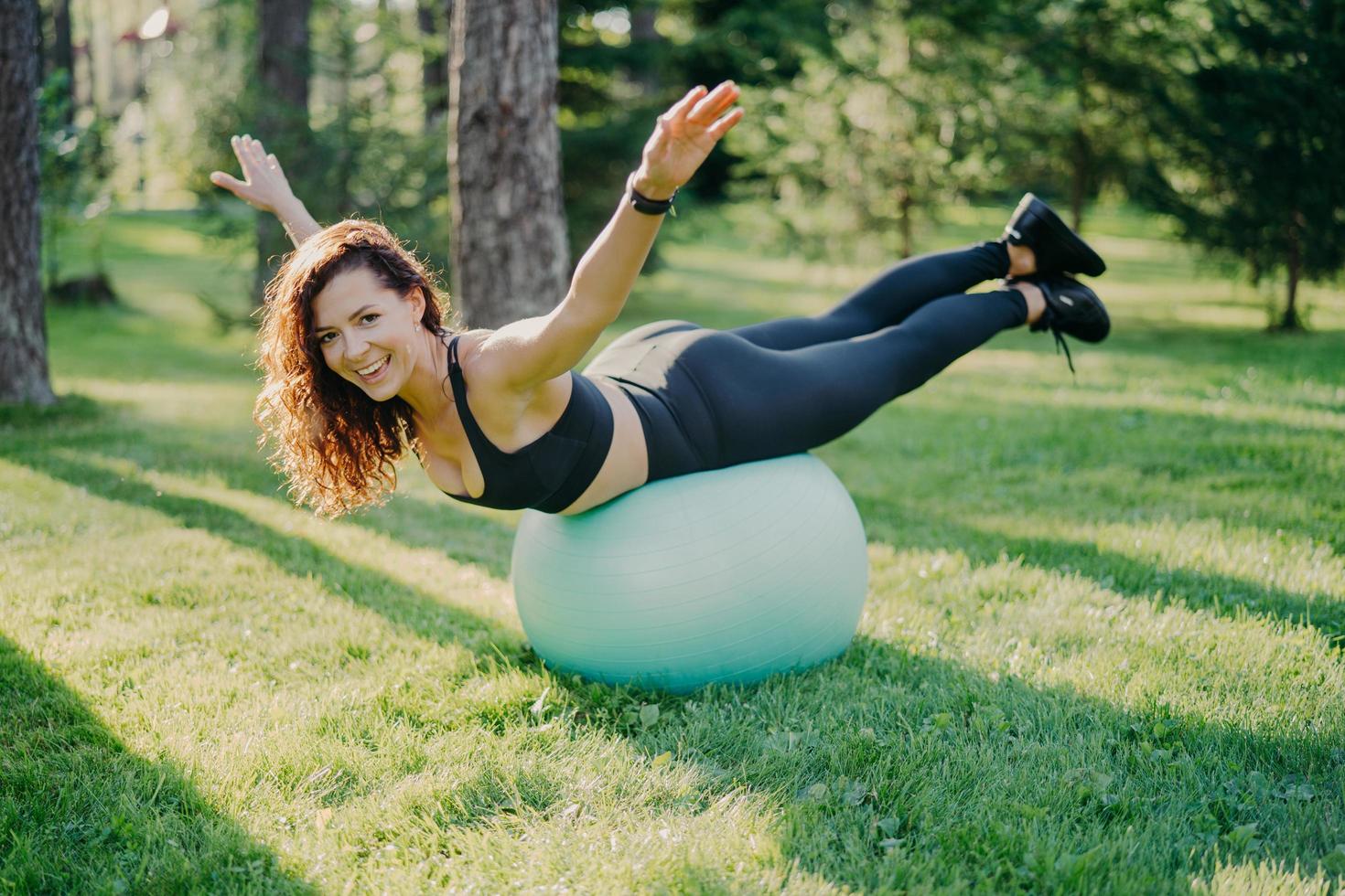 zufriedene fitte Frau trainiert im Freien auf Fitnessball hebt die Arme und versucht, in aktiver Kleidung gekleidet auszugleichen, hat einen perfekten gesunden Körper und macht Yoga-Übungen im Freien auf grünem Gras. Gesunder sportlicher Lebensstil foto