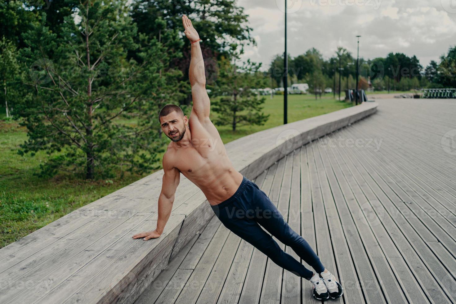 athletischer mann steht in seitlicher plank-pose, hebt einen arm, posiert mit hemdlosem oberkörper, trägt hose und schleichen. Sportler trainiert im Park an der frischen Luft, hat einen perfekten Muskelkörper. gesunder Lebensstil foto