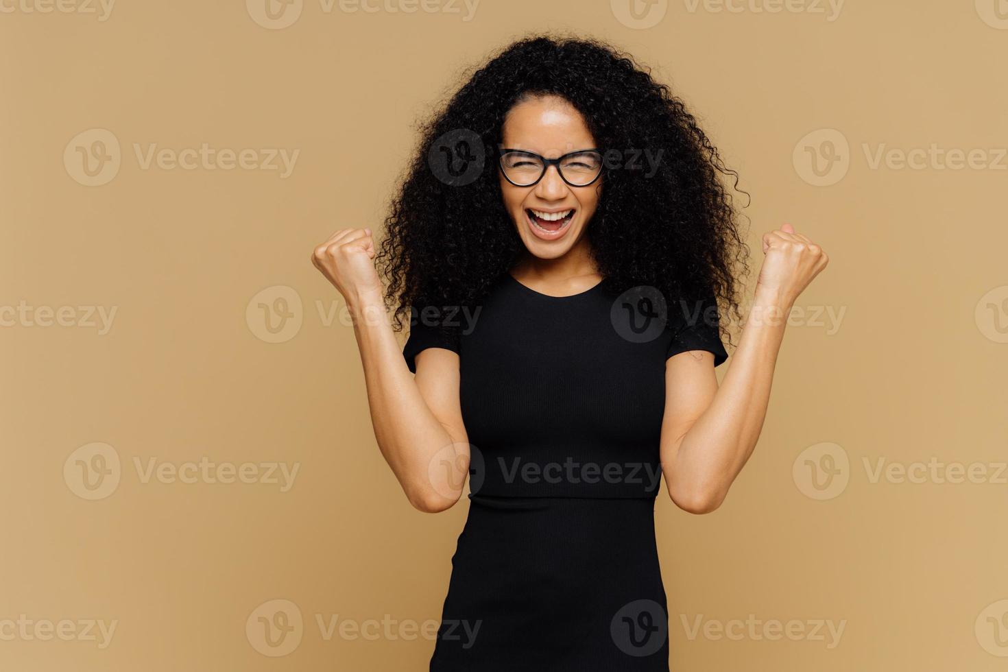 studioaufnahme einer überglücklichen jubelnden frau mit lockigem haar, hebt geballte fäuste, feiert erfolg und triumph, gekleidet in lässiges schwarzes t-shirt, brille, steht an brauner wand mit leerem raum foto