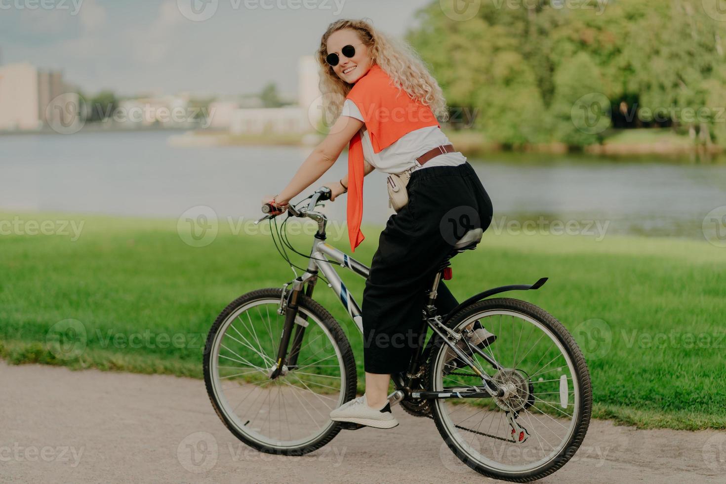 Zufriedene junge Frau genießt neue Route mit dem Fahrrad, fährt zwischen See, grünem Rasen und weit entfernten Gebäuden, trägt Sommersonnenbrillen, lässiges Outfit, weiße Turnschuhe, ist in guter körperlicher Verfassung foto