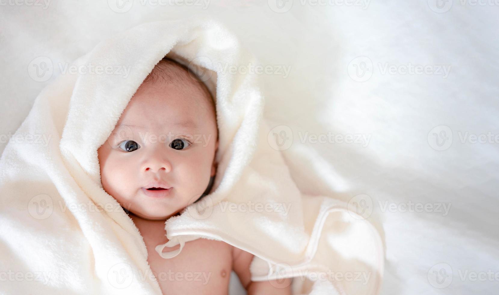 entzückendes, asiatisches neugeborenes, das mit weißer decke auf weißem bett im schlafzimmer liegt. kleiner Junge, der mit glücklichem Gesicht in die Kamera schaut. kleines unschuldiges neugeborenes kind am ersten lebenstag. Muttertagskonzept. foto