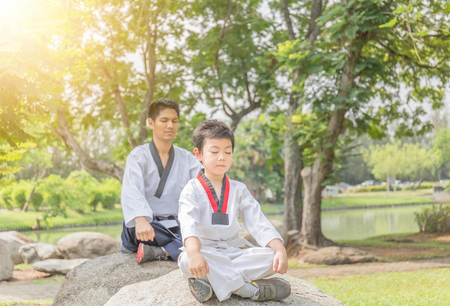 Taekwondo-Mann und Kind, die im Park auf Stein sitzen und meditieren foto