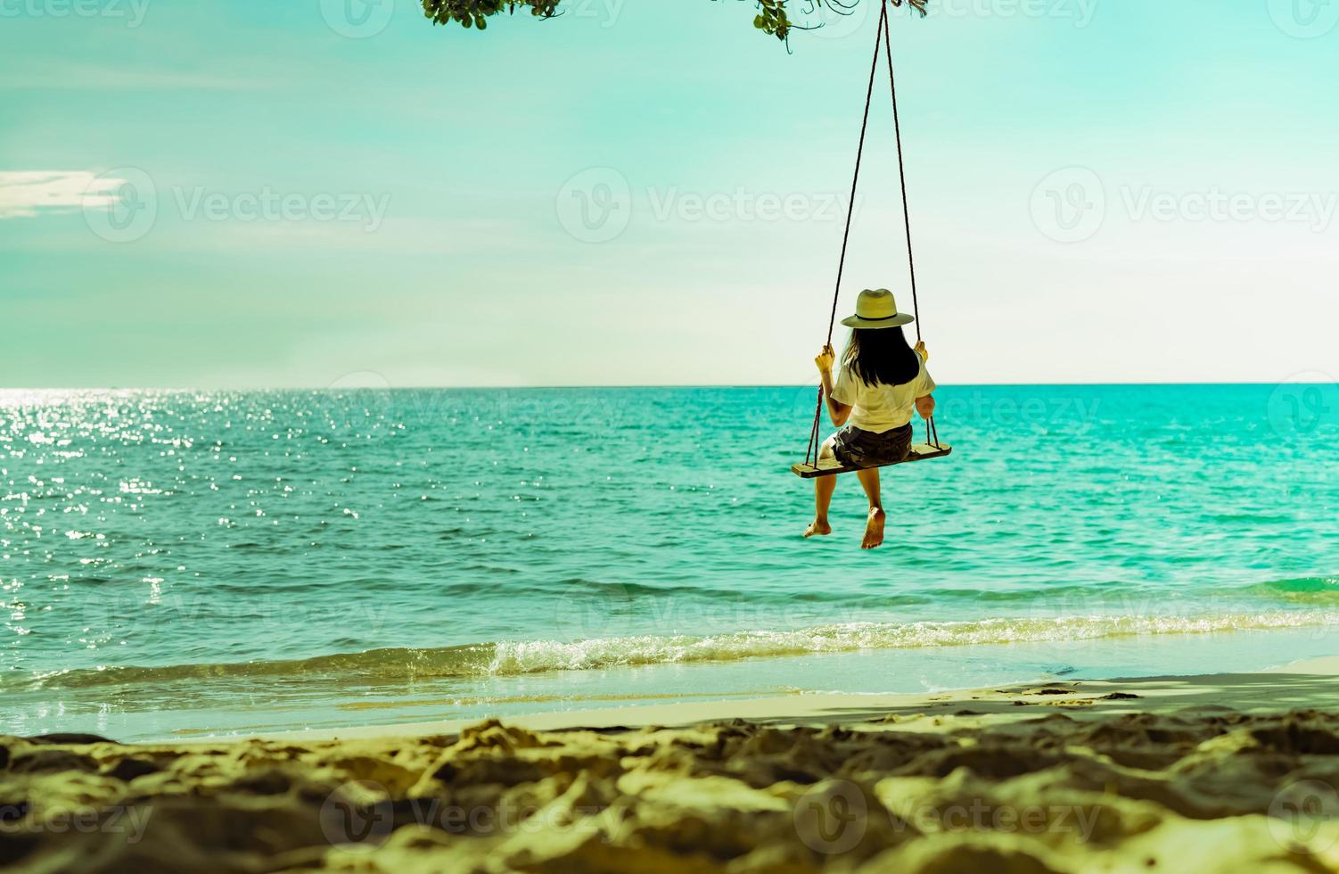 asiatische frau im lässigen stil trägt hut, schwingt die schaukeln am sandstrand und sieht bei sonnenuntergang ein wunderschönes tropisches paradies meer und himmel aus. Sommerurlaub. sommergefühl. genießendes und entspannendes Mädchen im Urlaub foto