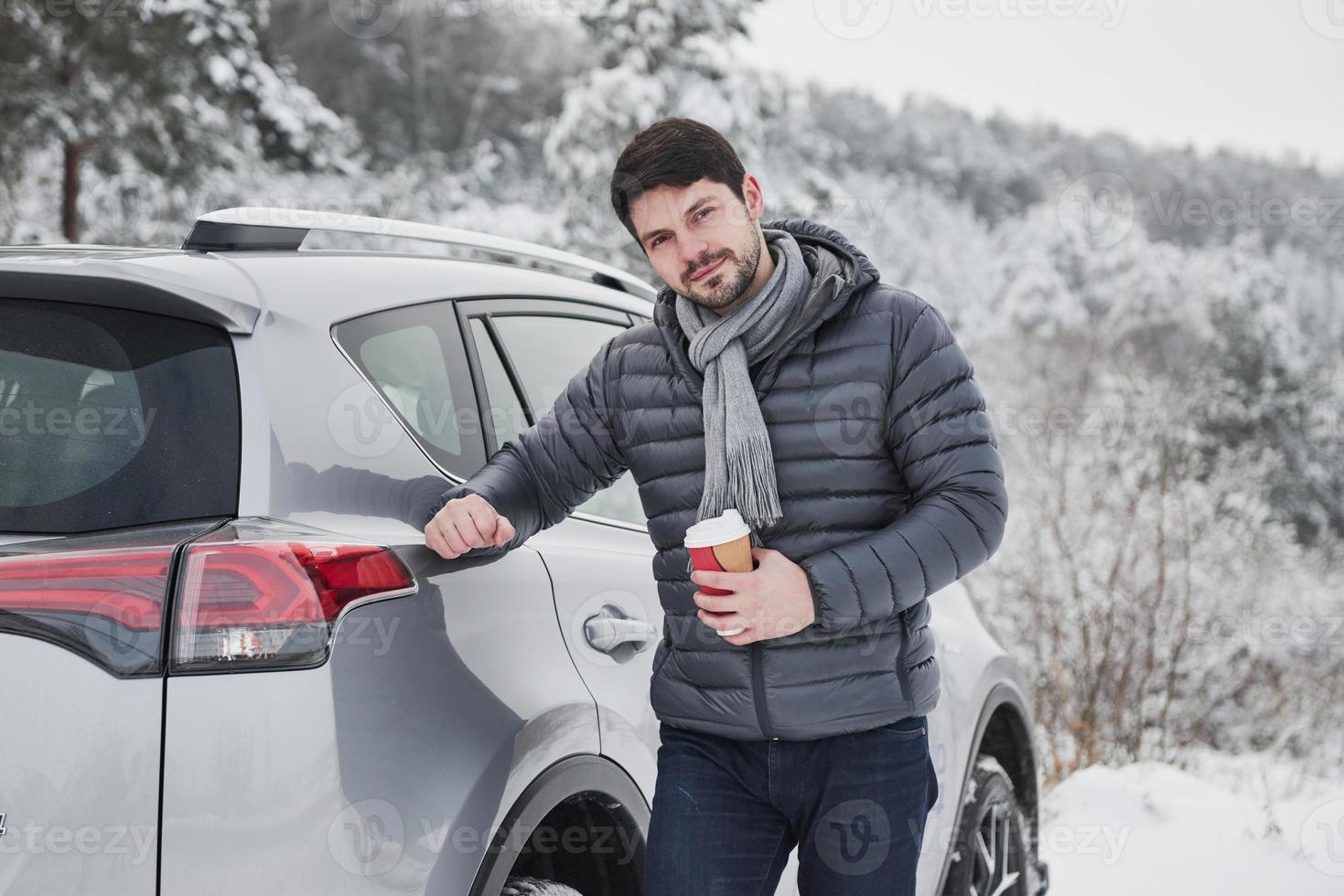 porträt des autobesitzers, der an einem verschneiten wintertag mit einer tasse kaffee in der hand in der nähe seines autos steht foto