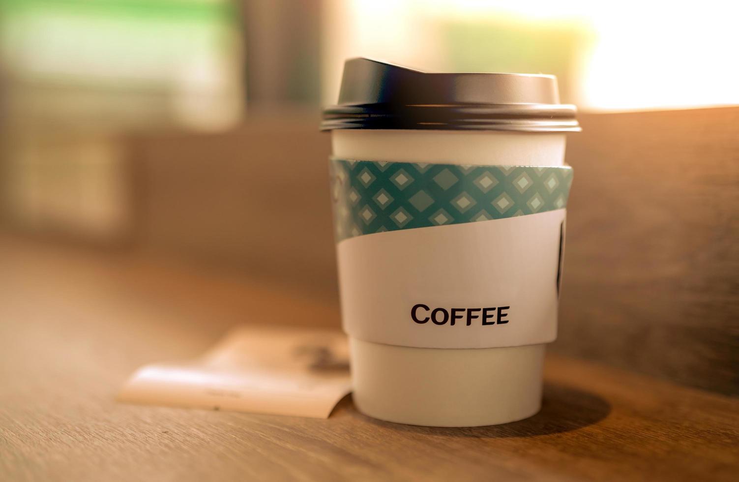 Kaffeeglas aus Papier im Café auf braunem Holztisch im Café am Morgen mit Sonnenlicht. Latte, Cappuccino oder Espresso im Papierglas. Einweg-Pappbecher. grüne Verpackung. heisses Getränk. foto