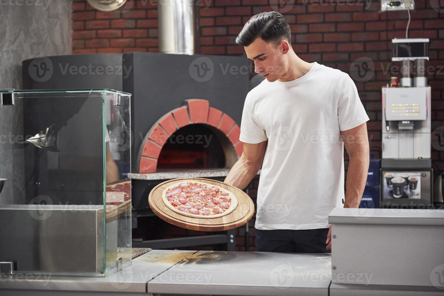 am Arbeitsplatz. Bäcker im weißen Hemd mit Pizza, die zum Kochen in den Ofen gestellt werden kann foto