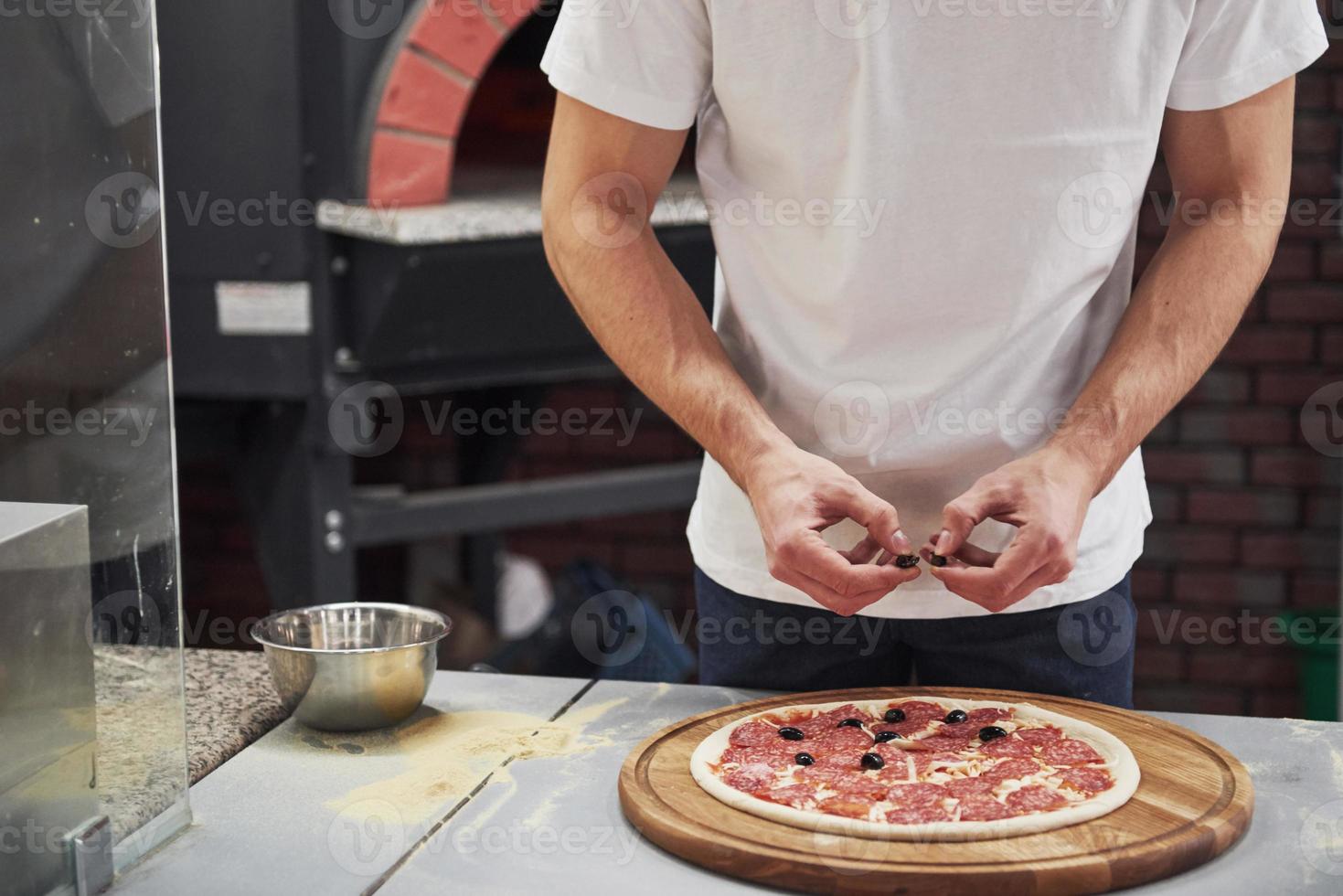köstliches Produkt herstellen. Bäcker im weißen Hemd, der Oliven legt, um köstliche Pizza für eine Bestellung im Restaurant zuzubereiten foto