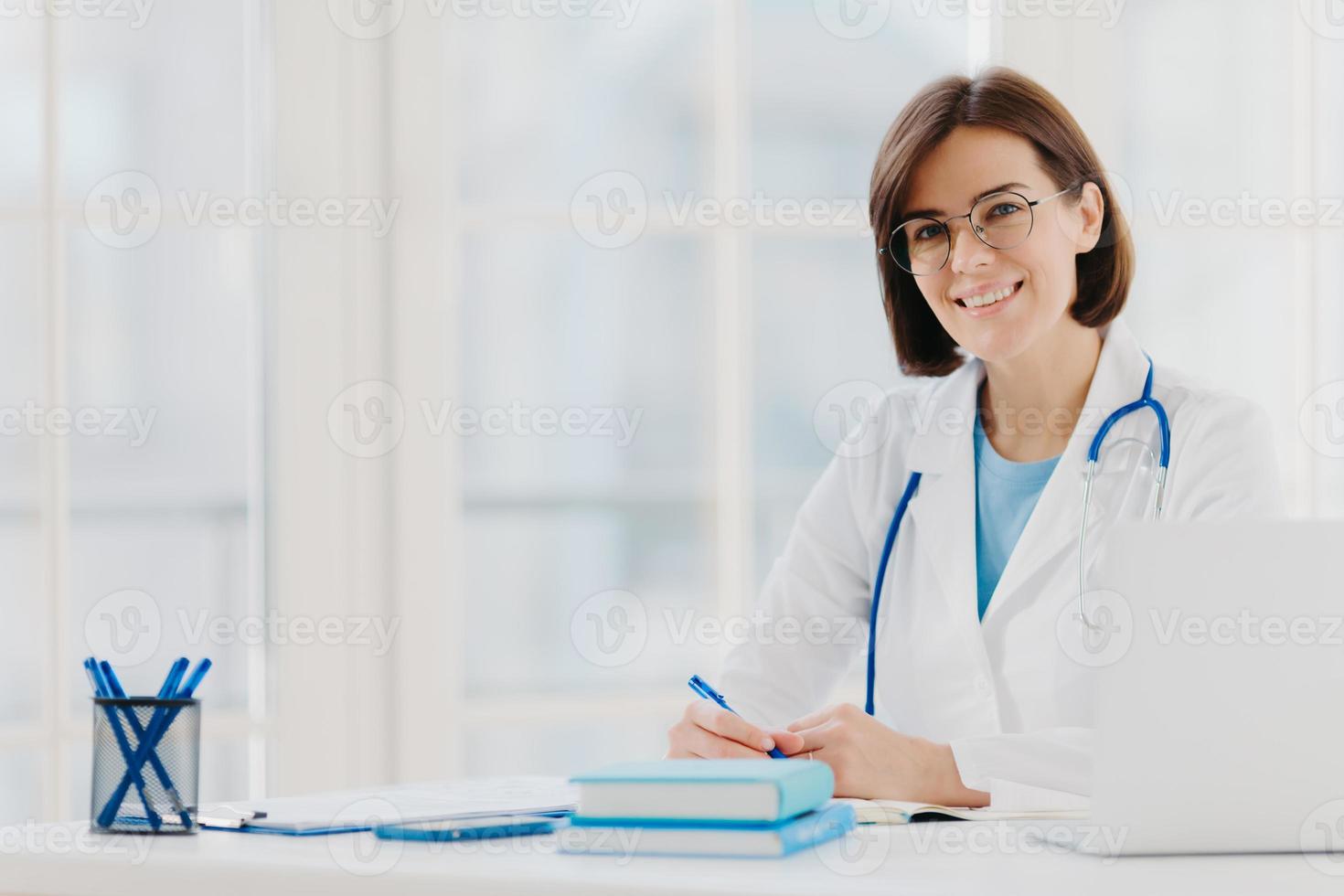 professionelle ärztin schreibt notizen auf, posiert mit laptop am schreibtisch im büro, trägt weißen kittel, brille und phonendoskop um den hals, schaut durch medizinische dokumente. Gesundheitskonzept foto