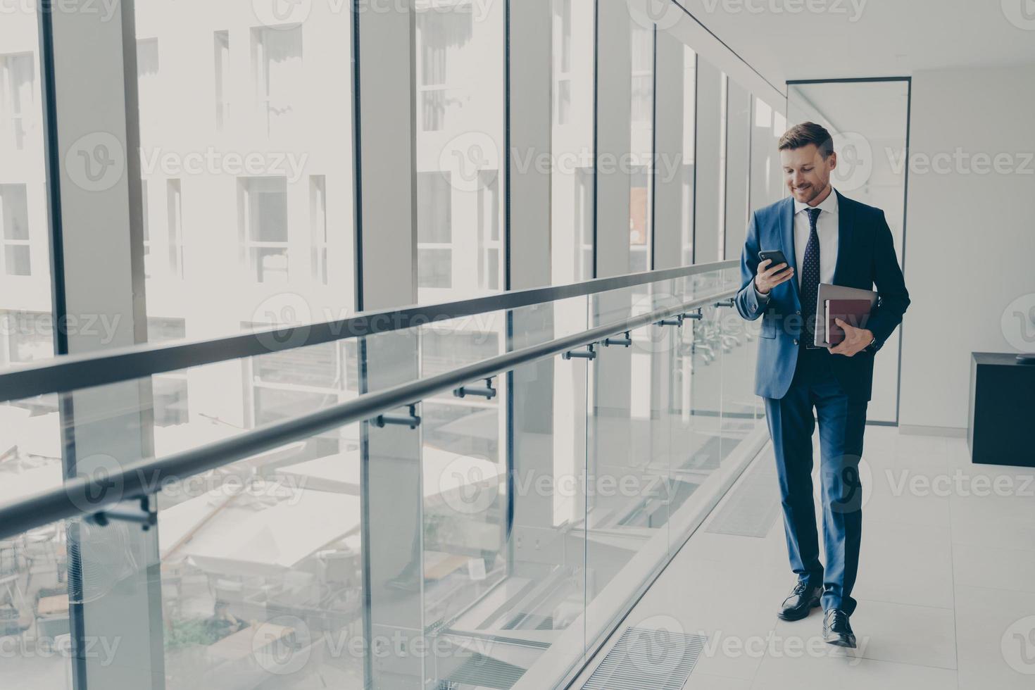 lächelnder rothaariger Büroangestellter im Anzug mit Handy, während er im modernen Büro steht foto