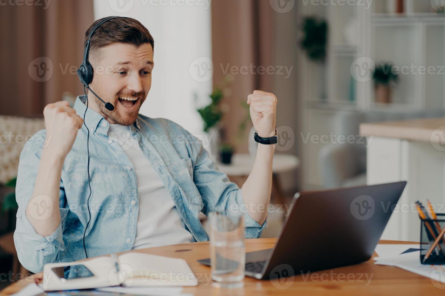 Überglücklicher, triumphierender erwachsener Mann ballt die Fäuste, feiert den Erfolg, schaut aufmerksam auf den Laptop-Bildschirm, trägt ein Headset, hat Firmenmeeting-Online-Posen auf dem Desktop vor verschwommenem Hintergrund foto