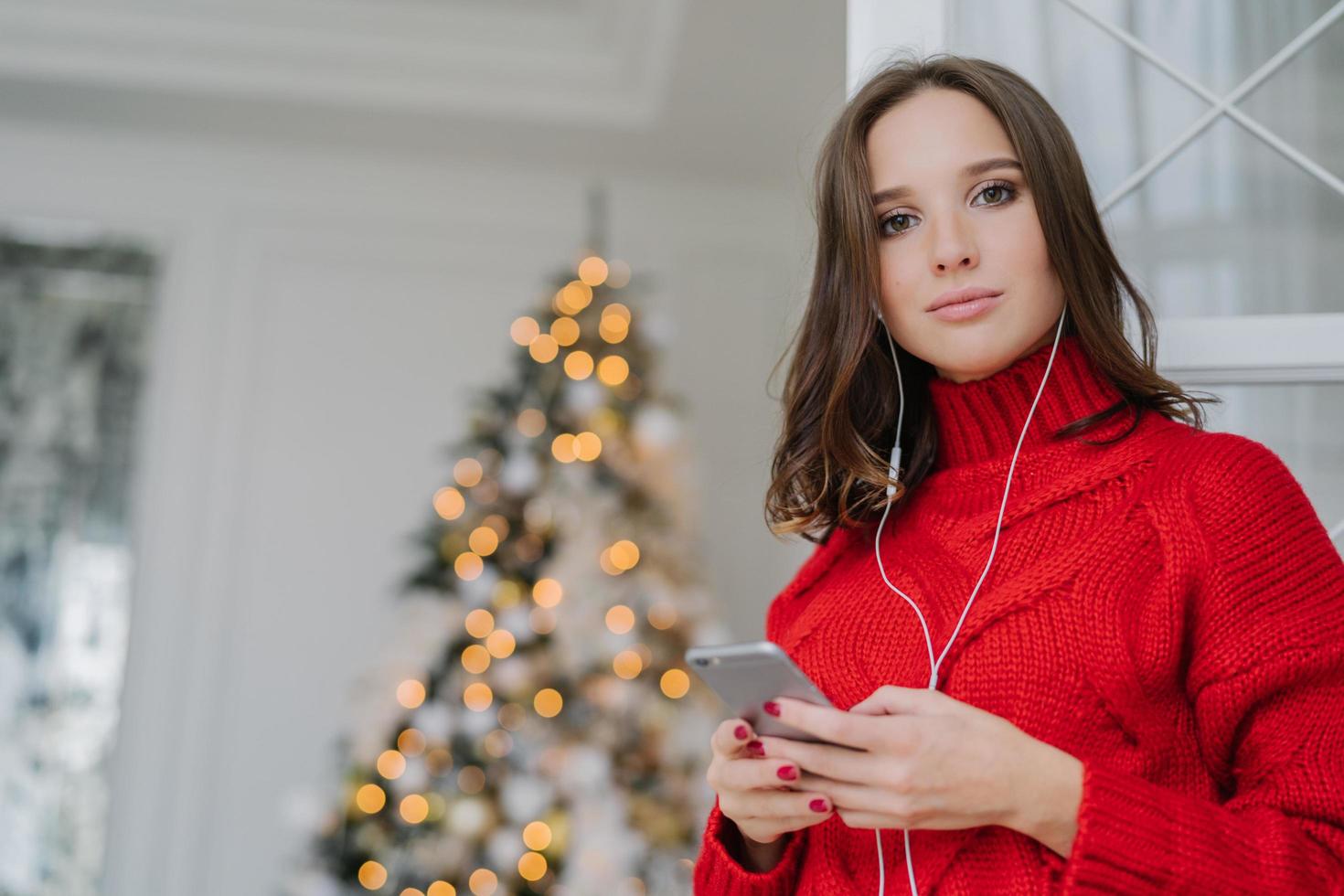 foto einer gutaussehenden frau mit dunklem haar, gekleidet in einen roten strickpullover, surft in sozialen netzwerken auf dem handy, hört musik mit kopfhörern, bewundert den geschmückten weihnachtsbaum. Freizeitkonzept