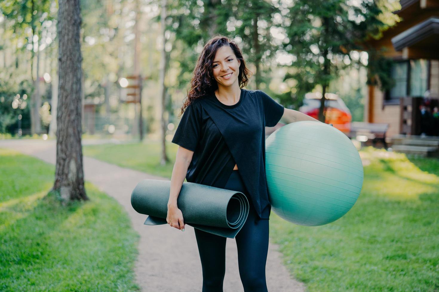 frohe brünette junge frau lächelt positiv hält großen fitnessball und karemat, gekleidet in schwarzes t-shirt, leggings, macht gymnastikübungen im freien, hat perfekte figur, gesunden körper foto