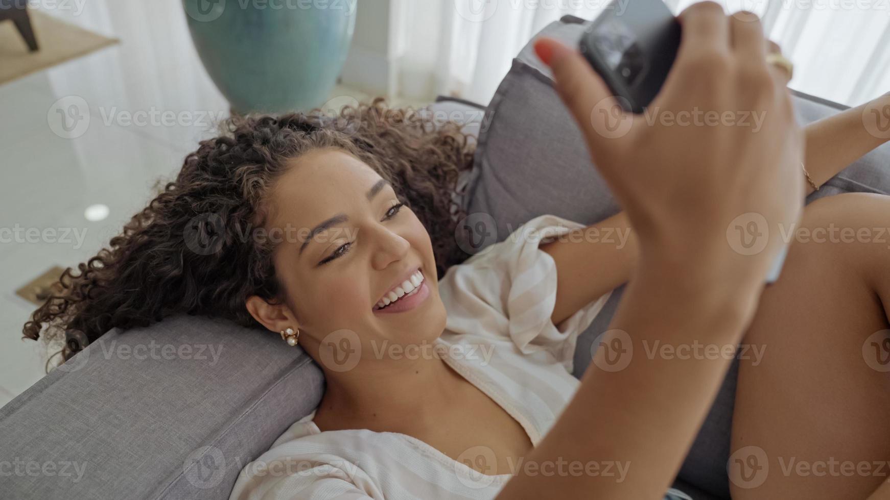 glückliche lateinische dame entspannen sich allein zu hause sitzen in der zimmerpose teilen gute nachrichten in den sozialen medien über das handy. foto